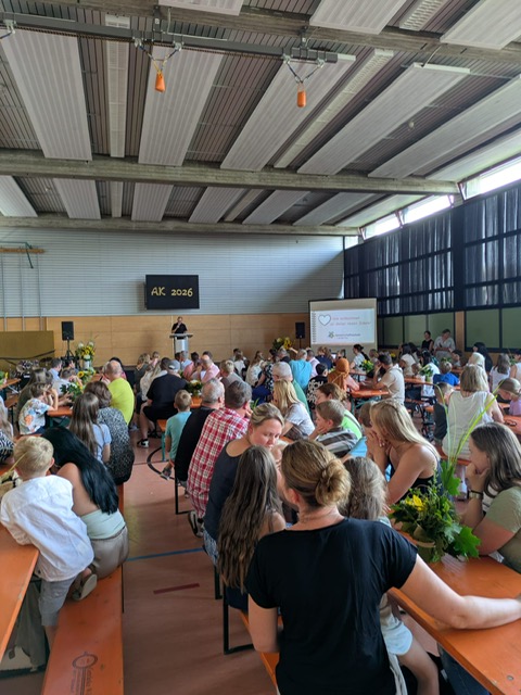 Viele Gäste sitzen auf Bierbänken in der Turnhalle vor der Bühne. Jemand hält eine Rede.