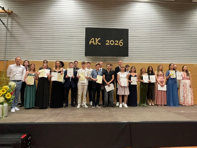 Schülerinnen und Schüler der Klasse 10a stehen mit schicker Kleidung auf der Bühne und präsentieren ihre Abschlusszeugnisse mit den Schulleitungen und der Klassenlehrkraft