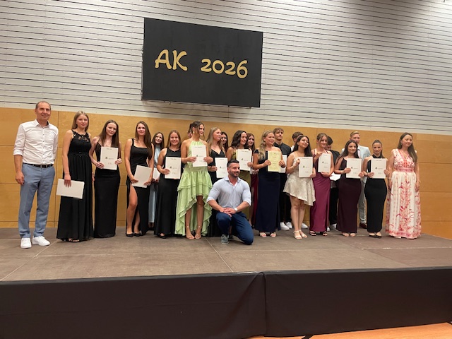 Schülerinnen und Schüler der Klasse 10b stehen mit schicker Kleidung auf der Bühne und präsentieren ihre Abschlusszeugnisse mit den Schulleitungen und der Klassenlehrkraft