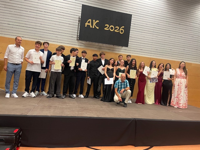 Schülerinnen und Schüler einer neunten Klasse stehen mit schicker Kleidung auf der Bühne und präsentieren ihre Abschlusszeugnisse mit den Schulleitungen und der Klassenlehrkraft